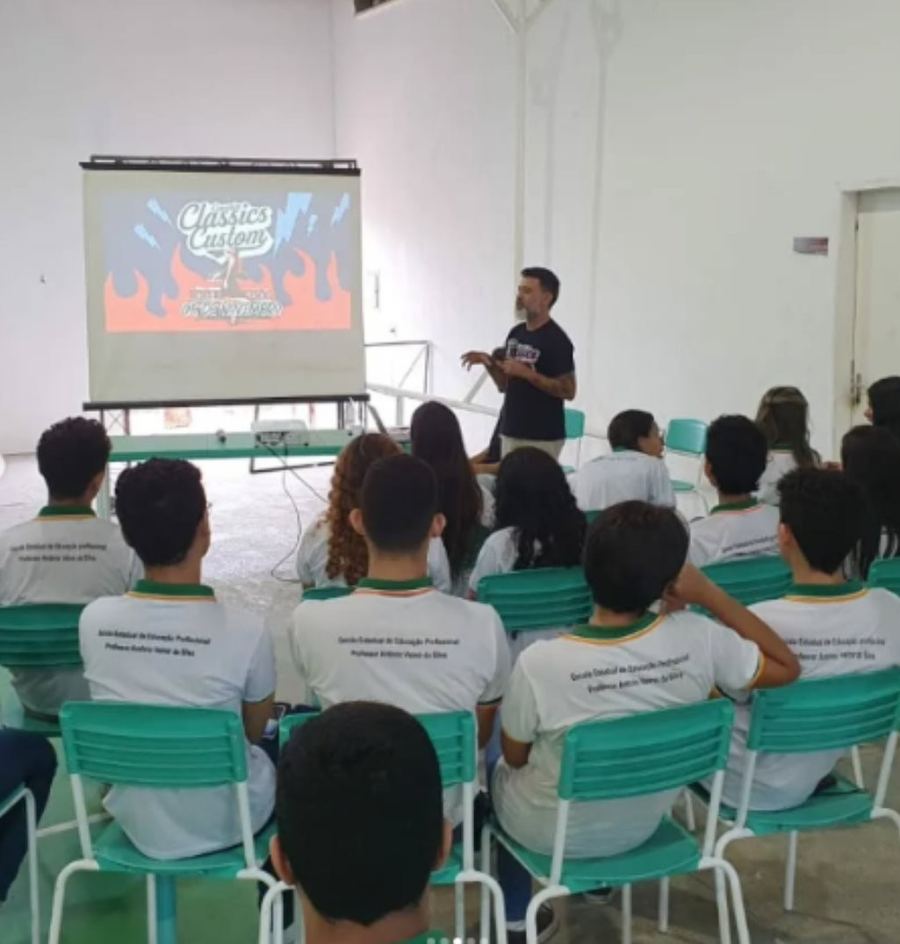 Estudantes de uniforme sentados em uma sala assistindo a uma palestra projetada na parede