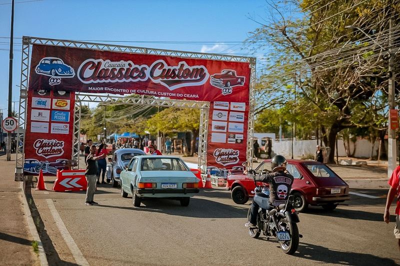 Entrada do Caucaia Classics Custom com carros antigos e uma moto passando sob o arco do evento