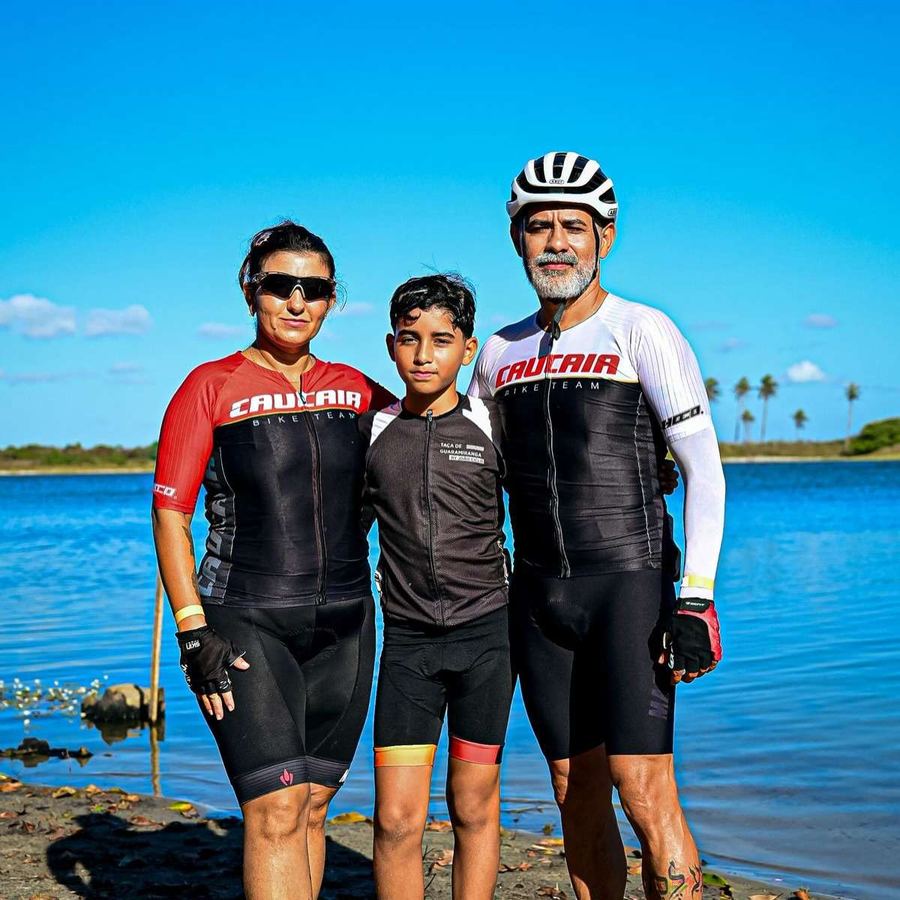 Três ciclistas de uniforme posam diante de uma lagoa em dia de sol